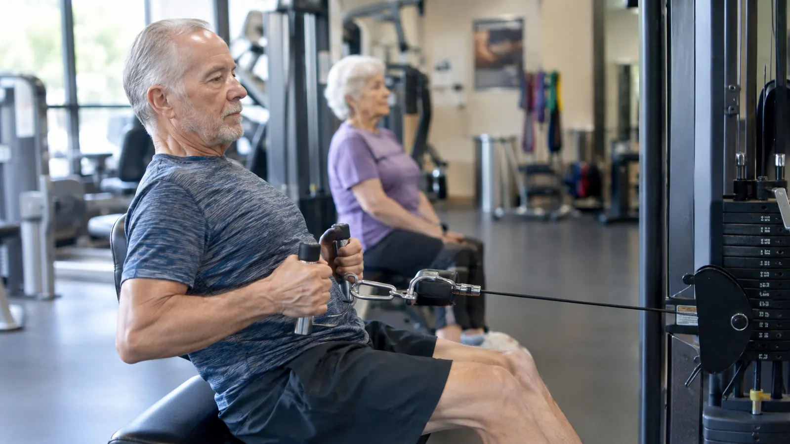 An older man in his sixties doing seated rows on a cable machine at a rec center, with another older adult nearby